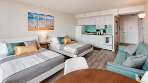 A compact hotel room with two beds, a kitchenette, and a seating area.