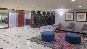 A lobby with a tiled floor, a stone wall with plants, wicker chairs, and a colorful rug.