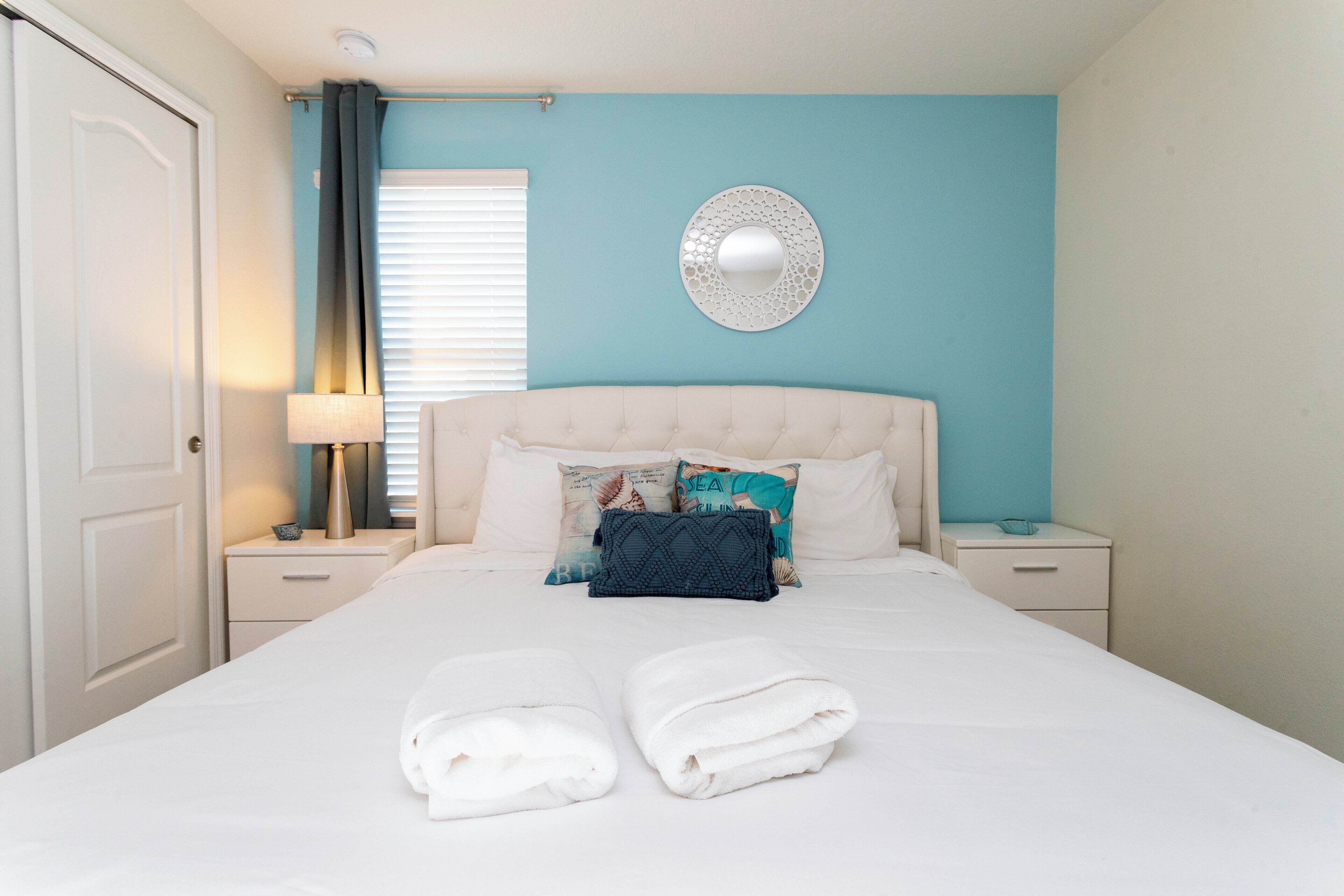 A neatly made bed with two folded towels, a bedside lamp, and a decorative mirror on the wall.