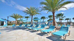 Un espace aménagé au bord de la piscine, avec des chaises longues bleues et des parasols, des palmiers et un ciel d’un bleu limpide.