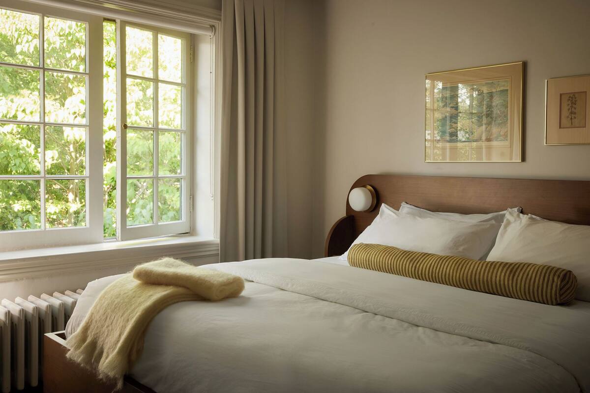 Un lit bien fait, avec une couette blanche et un oreiller à rayures, une fenêtre avec des rideaux et deux tableaux encadrés accrochés au mur.