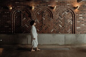Una persona caminando frente a un muro de ladrillo con aberturas arqueadas y luces fijadas en la pared.