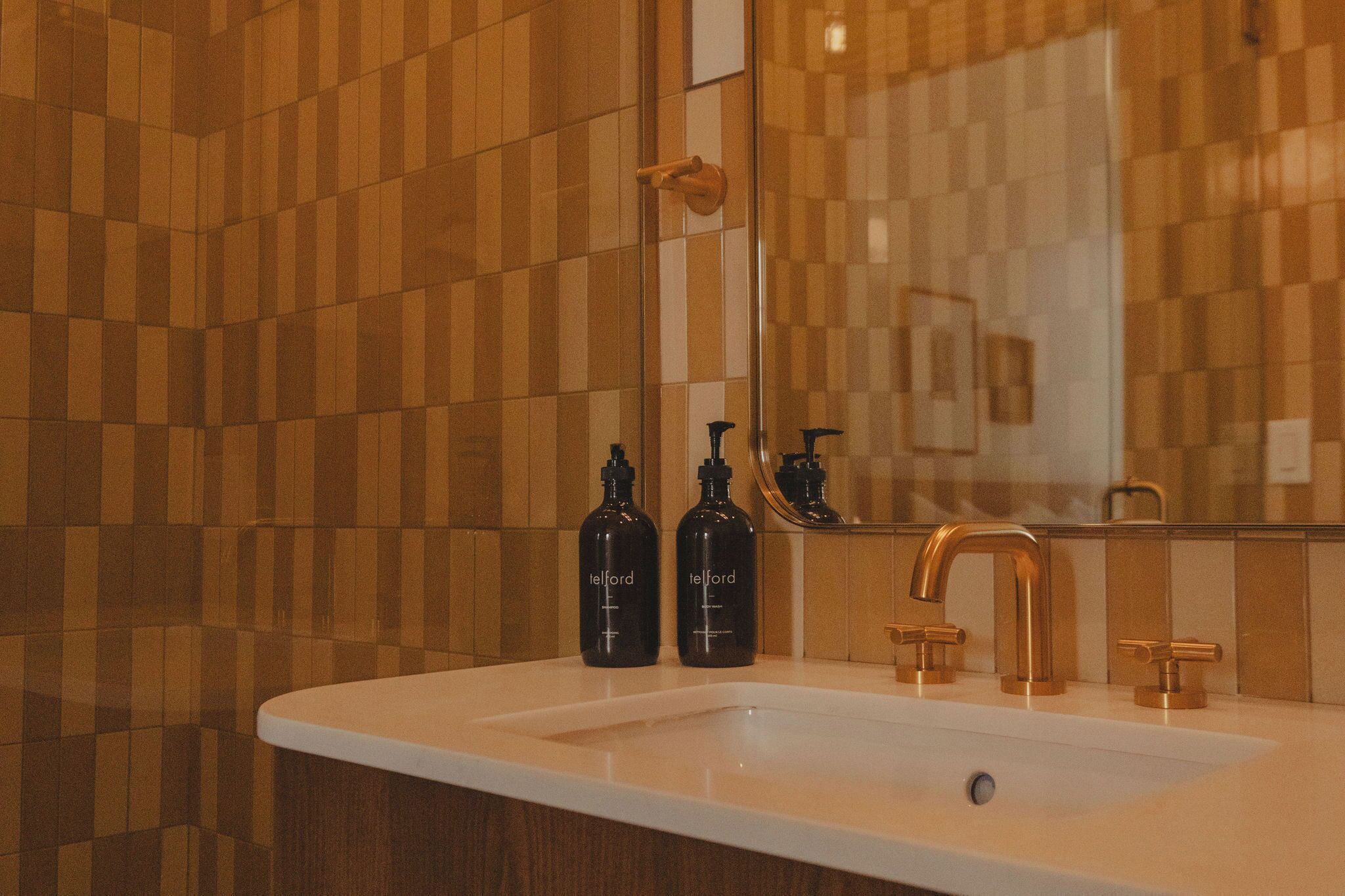 A bathroom with a white sink, two black bottles, a gold faucet, and a mirror reflecting a towel rack.
