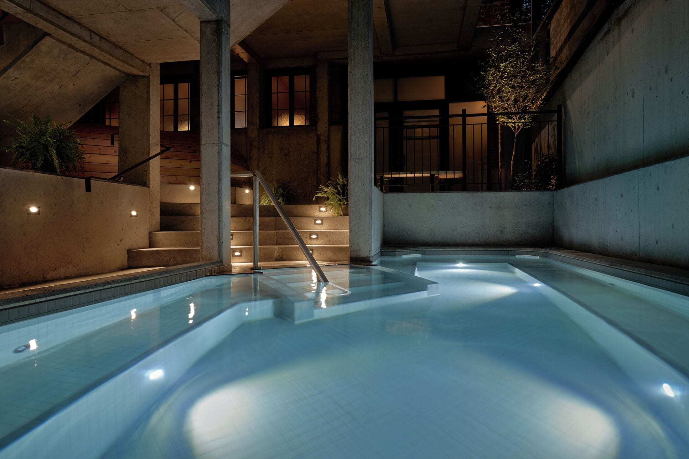 Una piscina amb una escala que condueix a un nivell superior, envoltada de murs de formigó i plantes en testos.