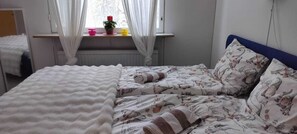 A bedroom with a bed, a window with curtains, a radiator, and a small table with decorative items. - LT Villa (Rovaniemi)
