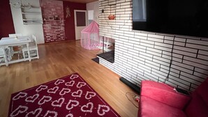 A living room with a TV mounted on a brick wall, a pink chair, a white table with chairs, and a red rug. - LT Villa (Rovaniemi)