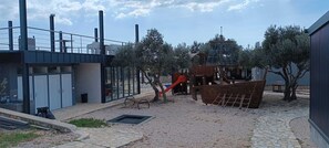 Explore the outdoor play area with a wooden slide and climbing structure, surrounded by trees and a gravel path.