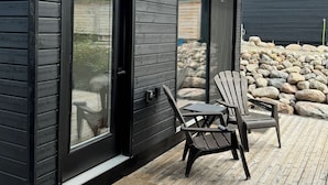 Ein modernes Haus mit dunkler Fassade, großen Glasüren und einer Holzterrasse mit zwei Adirondack-Stühlen.