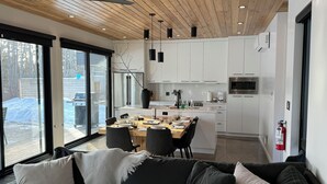Eine moderne Küche mit weißen Schränken, ein Essbereich mit einem Holztisch und schwarzen Stühlen sowie ein schwarzes Sofa mit weißen Kissen.