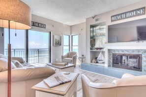 A spacious living room with a large TV, comfortable seating, a fireplace, and a view of the beach. - Oceanfront Paradise at Sea Fire Condos - 5B (New Smyrna Beach)