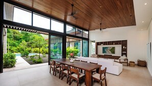 A spacious living area with a large wooden dining table, white sofa, and a TV mounted on the wall.
