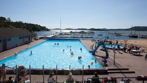 A swimming pool with multiple lanes, a water slide, and a view of boats and hills in the background. - Paradis på Sørlandet 2 (Agder)