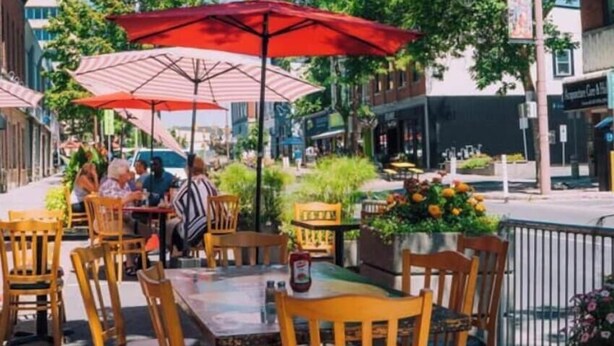 An outdoor cafe with tables and chairs under striped umbrellas, surrounded by greenery and buildings.