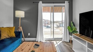Un salón moderno con un sofá azul, una mesa de centro de madera, una lámpara de pie, un televisor sobre un mueble de madera y un ventanal con cortinas.