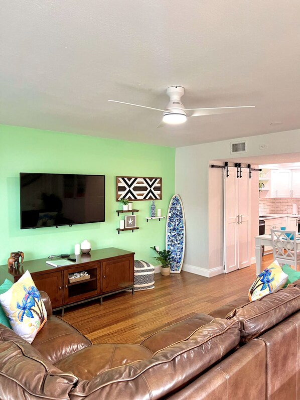 A living room with a brown leather sofa, a television, a ceiling fan, and a surfboard. - Steps to the Sand – 2BR Melbourne Beach Townhome (Melbourne Beach)