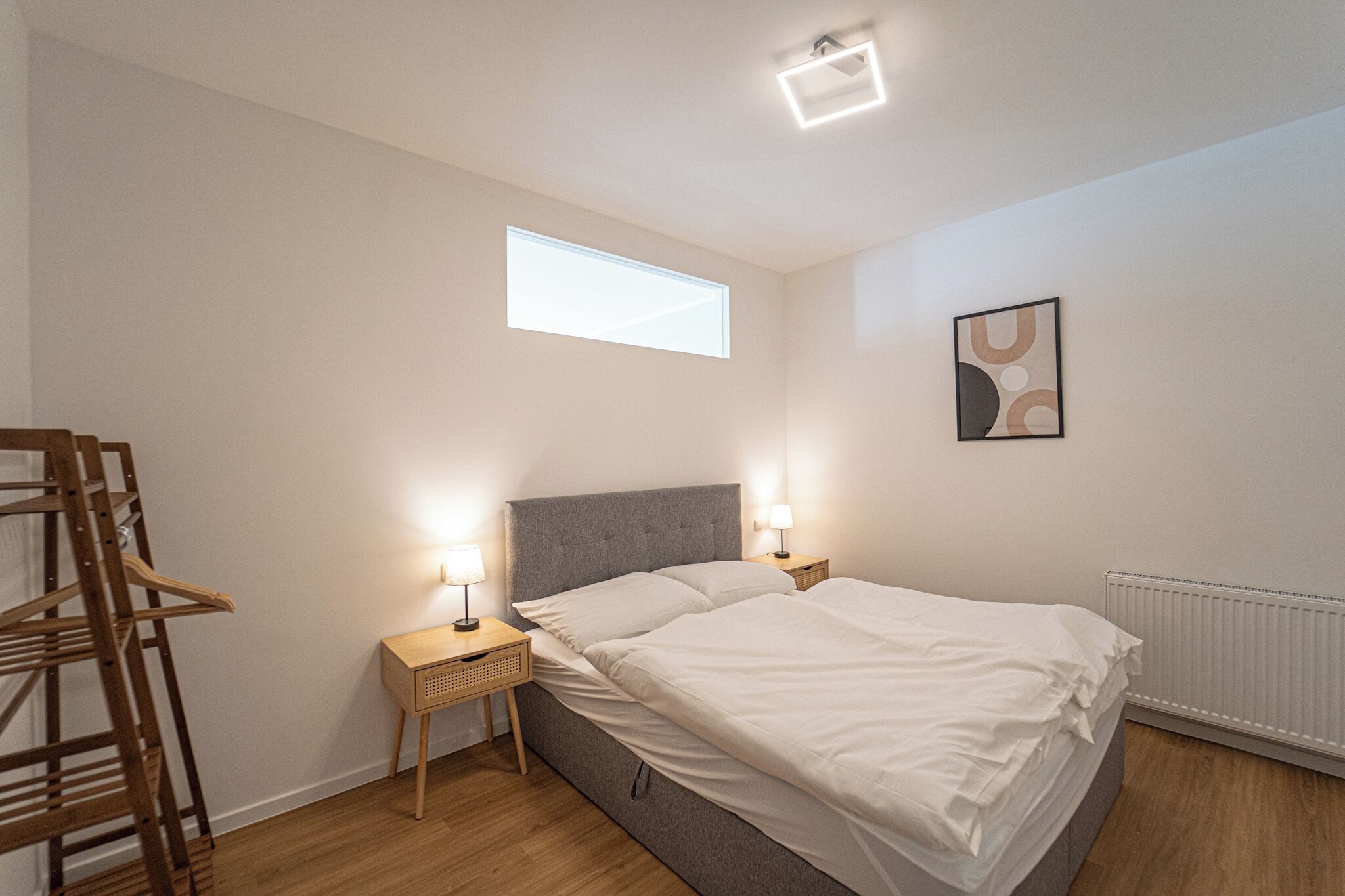 Un dormitorio con cama, lámparas de noche, una ventana, un radiador y una silla de madera.