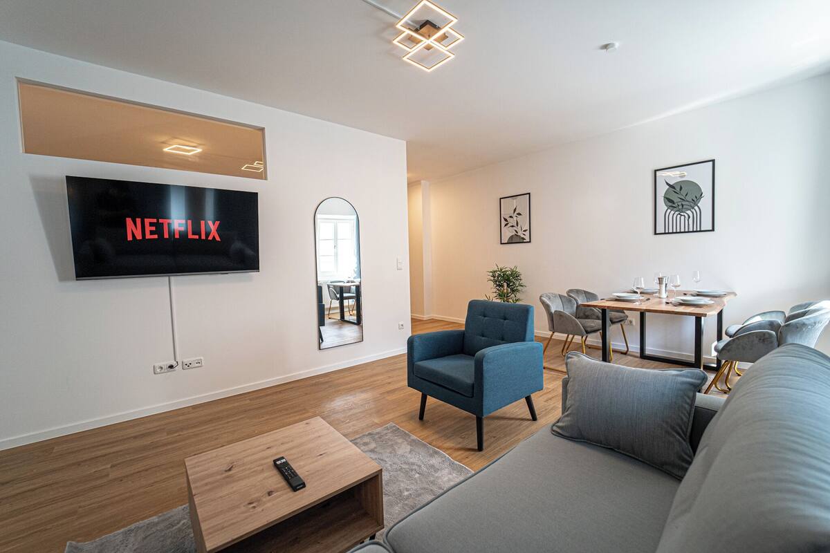 Een moderne woonkamer met een flatscreen televisie, een bank, een blauwe fauteuil, een houten salontafel en een eethoek met een tafel en stoelen.