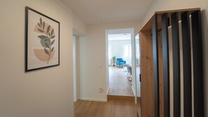Un couloir orné d’un tableau encadré au mur, un escalier avec des marches en bois et une vue sur une pièce où l’on aperçoit une chaise bleue et une plante.