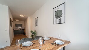 Un coin repas avec une table en bois dressée pour quatre personnes, une cuisine en arrière-plan et une œuvre d’art abstraite encadrée accrochée au mur.