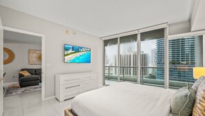 Un dormitorio moderno con una cama grande, un televisor en la pared, un balcón con vistas a la ciudad y un sofá gris en la esquina.