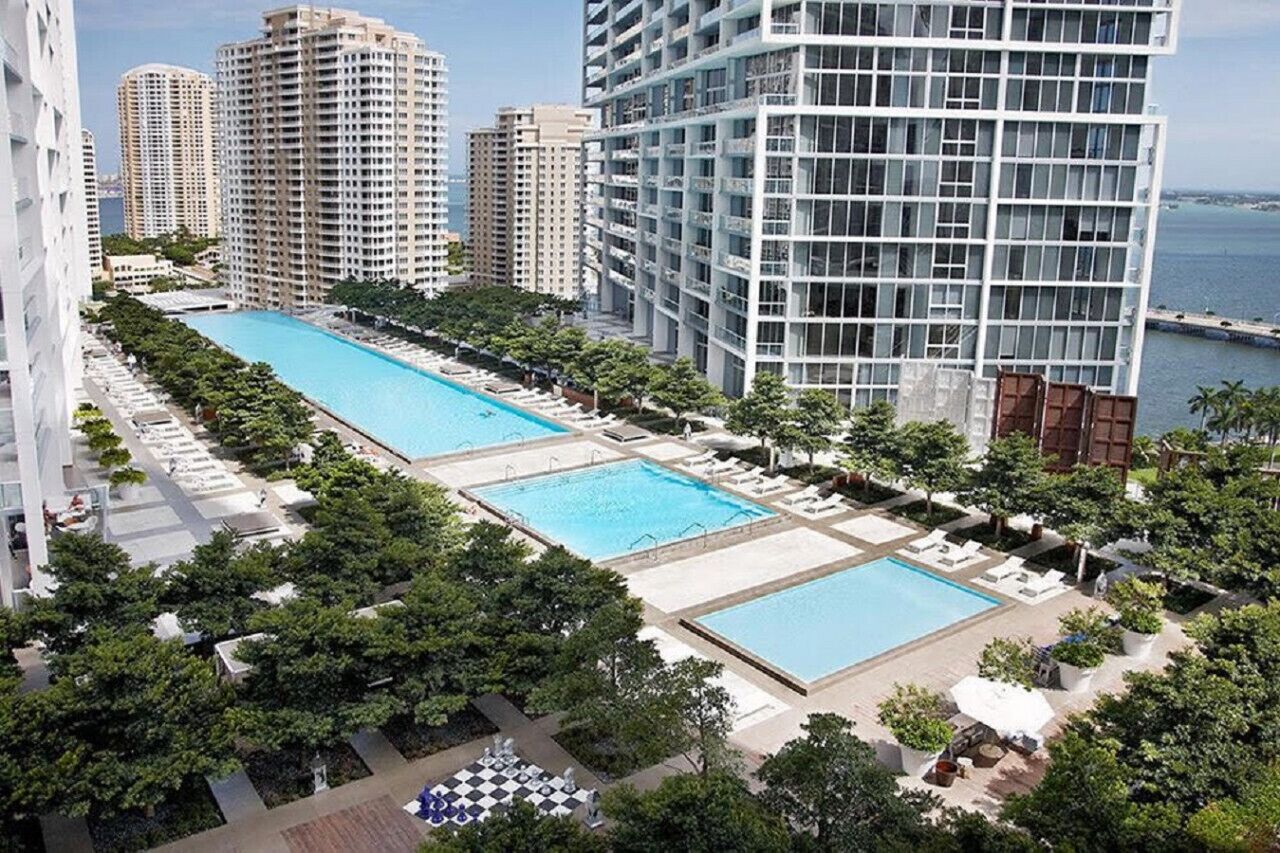 Un edificio alto con area piscina sul tetto, circondato da vegetazione lussureggiante e altri grattacieli.