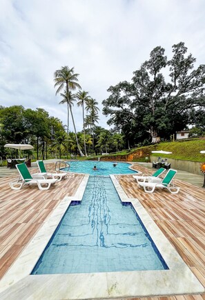 Piscina retangular com deck de madeira, rodeada por palmeiras e espreguiçadeiras.