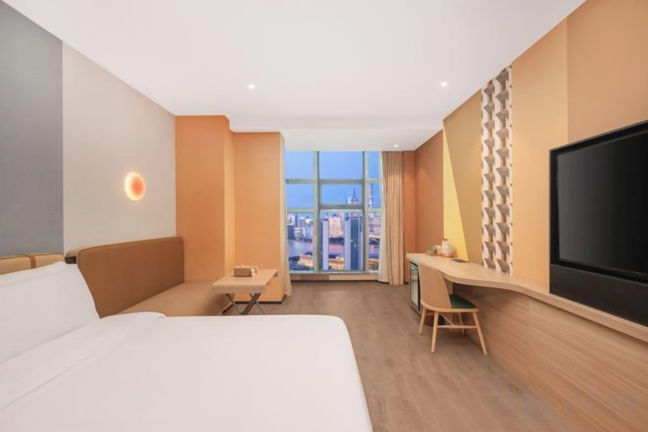 A hotel room with a bed, desk, chair, TV, and a view of the city.