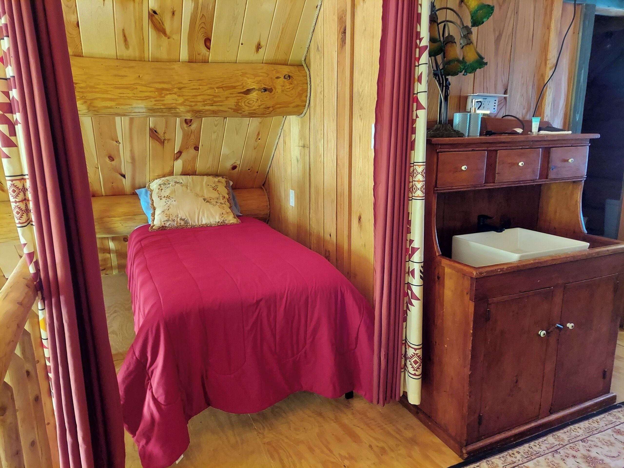 Une pièce de type cabane en bois comprenant un lit, un meuble en bois avec un évier et une fenêtre avec des rideaux.