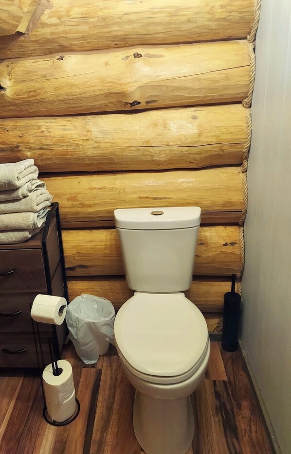 A wooden cabin-style bathroom with a toilet, a wooden cabinet with towels, and a roll of toilet paper.