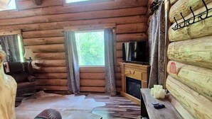 Interior de una cabaña de troncos con piso de madera, un sofá, un televisor y una chimenea.