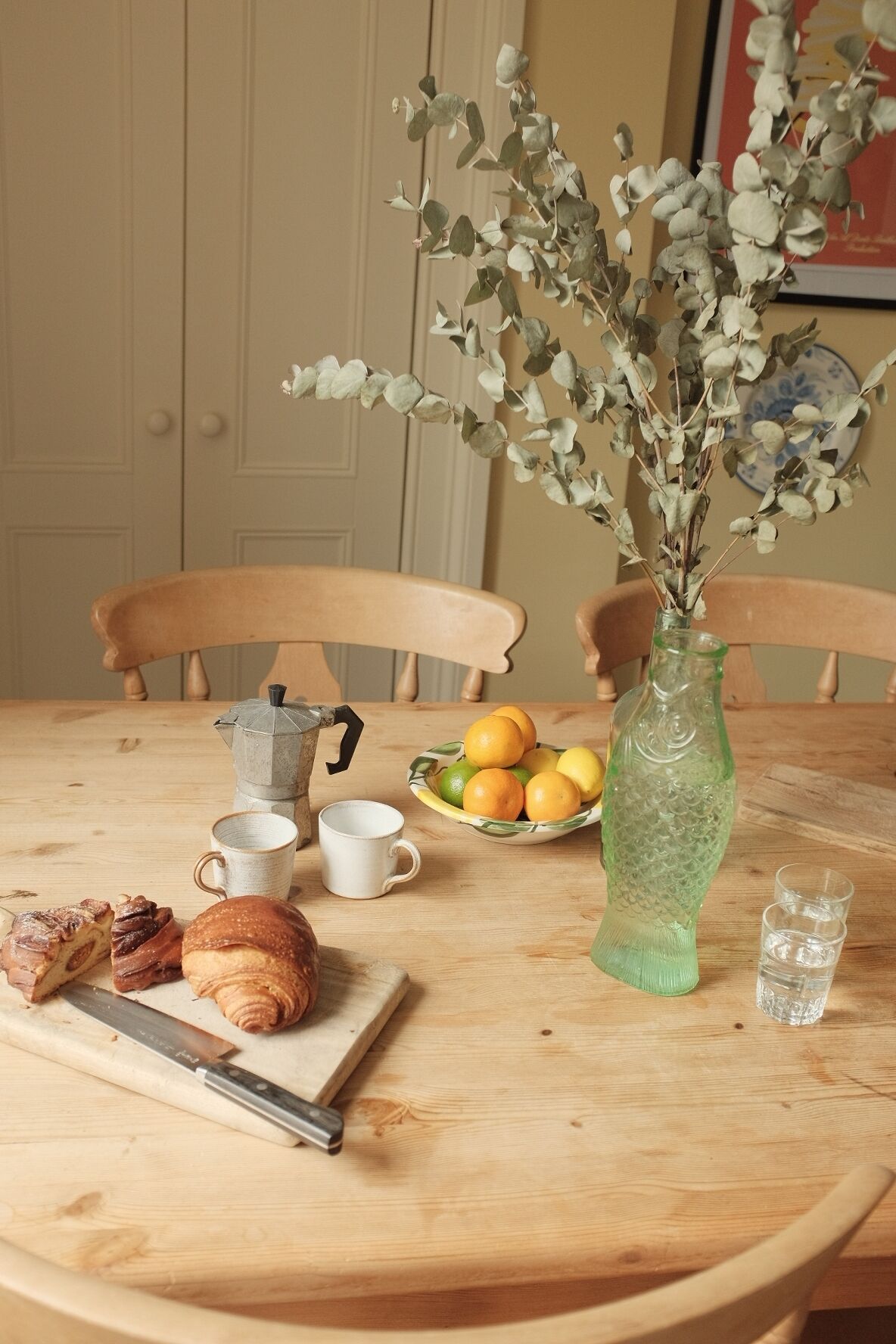 A wooden table with a green glass vase containing eucalyptus branches, a bowl of citrus fruits, a coffee pot, and a croissant.