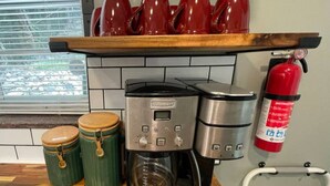 Une cuisine équipée d’une machine à café, de deux récipients verts, d’un extincteur rouge et d’une fenêtre avec des stores.