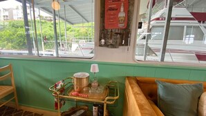 Une pièce avec un mur vert, un banc en bois, un chariot en métal contenant divers objets, et une fenêtre donnant sur un paysage verdoyant et un bateau.