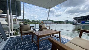 Un pont de bateau avec un plancher à motifs bleus, une table, deux chaises et une vue sur l’eau et les bâtiments éloignés.
