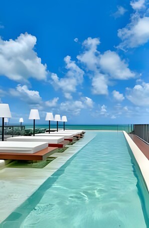 Zona de piscina con sillones y sombrillas, con vista a un mar azul cristalino.
