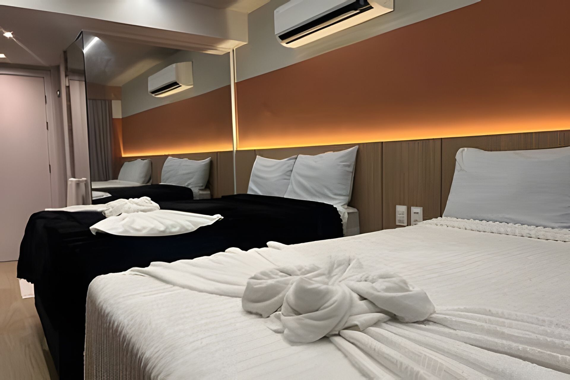 A hotel room with two beds, each with white linens and a towel. There are air conditioning units on the ceiling and a mirror on the wall.