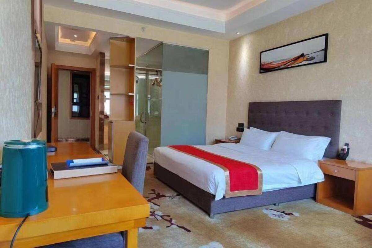A hotel room with a bed, bedside table, and a desk with a chair. There is a coat rack and a mirror.