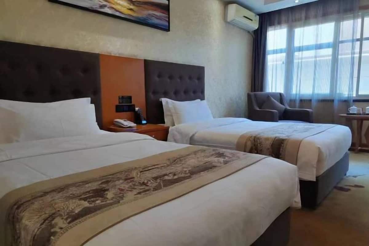 A hotel room with two beds, a headboard, a chair, a small table, and a window with curtains.