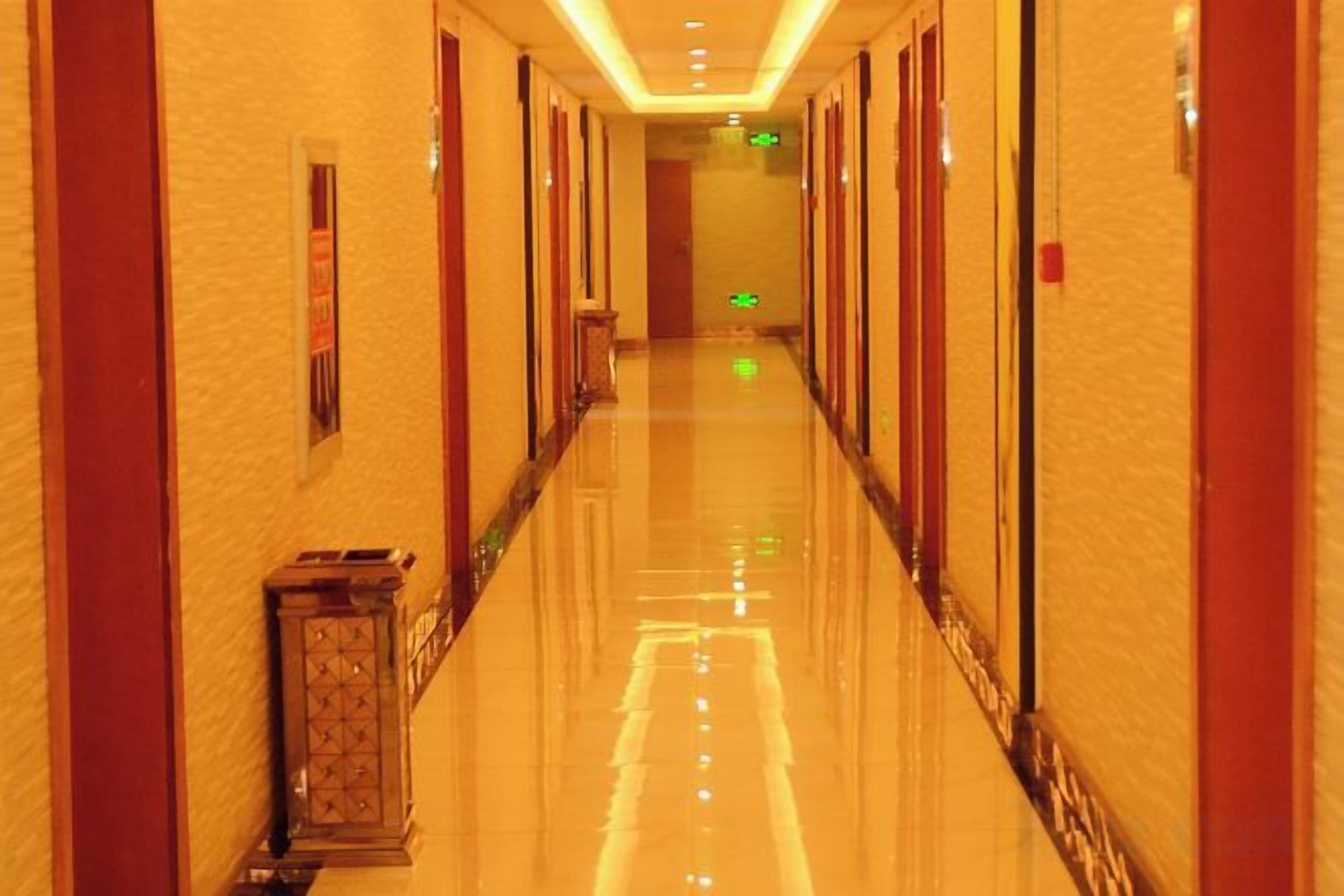 A hotel corridor with a polished floor, doors on both sides, and a green exit sign at the end.