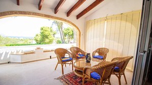 Une terrasse couverte, aménagée avec des meubles en osier, une table en verre, et offrant une vue sur la verdure et une route.