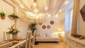 A cozy living room with a sofa, a dining table with chairs, a hanging light fixture, and a TV on the wall.
