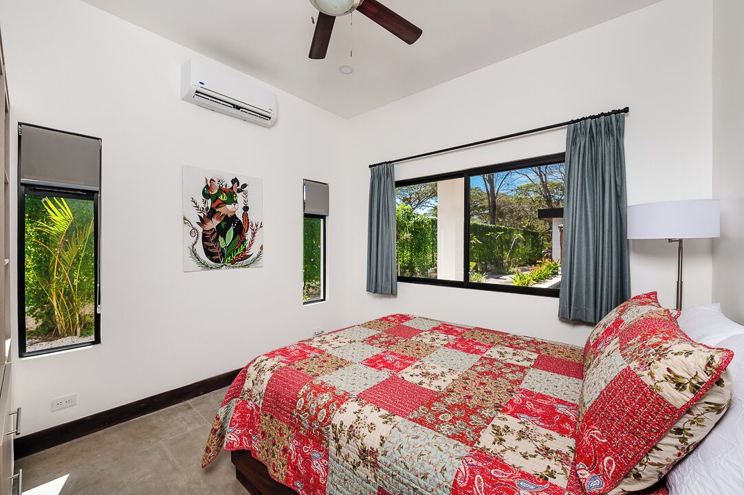 Un dormitorio con una cama grande, un ventilador de techo, un equipo de aire acondicionado, una ventana con cortinas y un televisor montado en la pared.