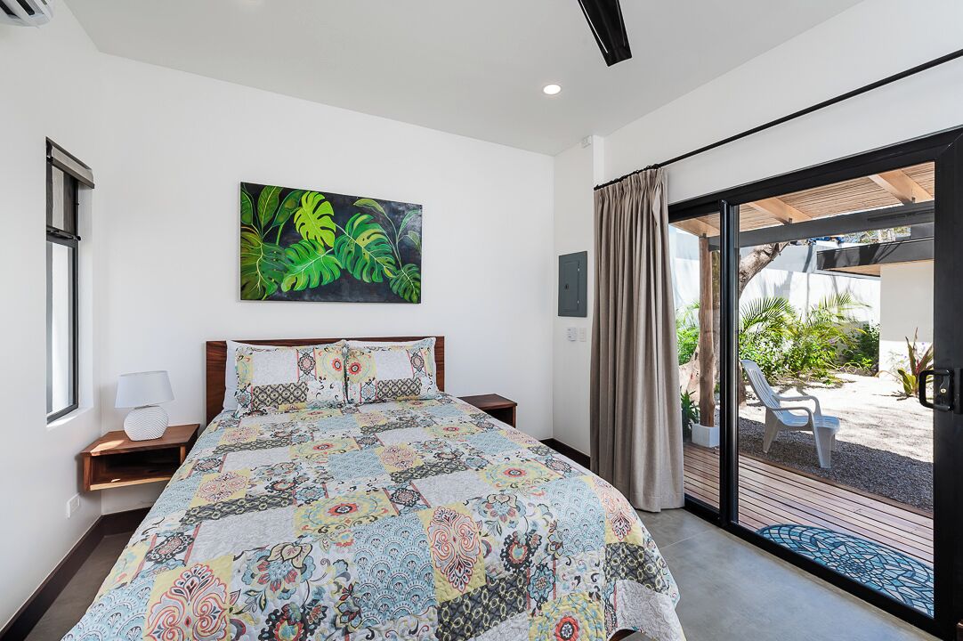 A bedroom with a large bed, a nightstand, a wall-mounted lamp, and a sliding glass door leading to an outdoor area with a patio and chairs.