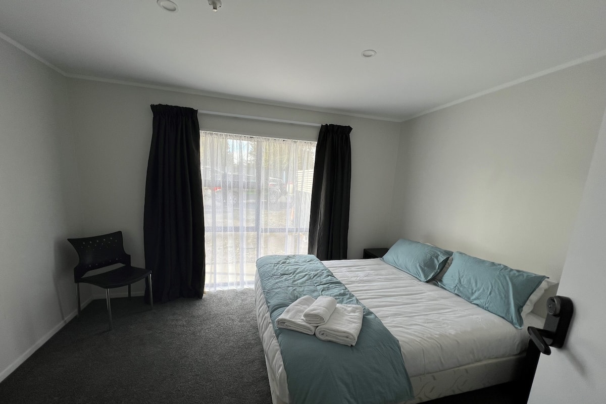 Quarto de hotel com cama, uma cadeira, mesa de cabeceira e janela com cortinas.