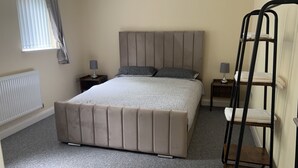 Ein Schlafzimmer mit einem großen Bett, zwei Nachttischen, einem Heizkörper, einem Fenster mit Vorhängen und einem Stuhl mit Rückenlehne.