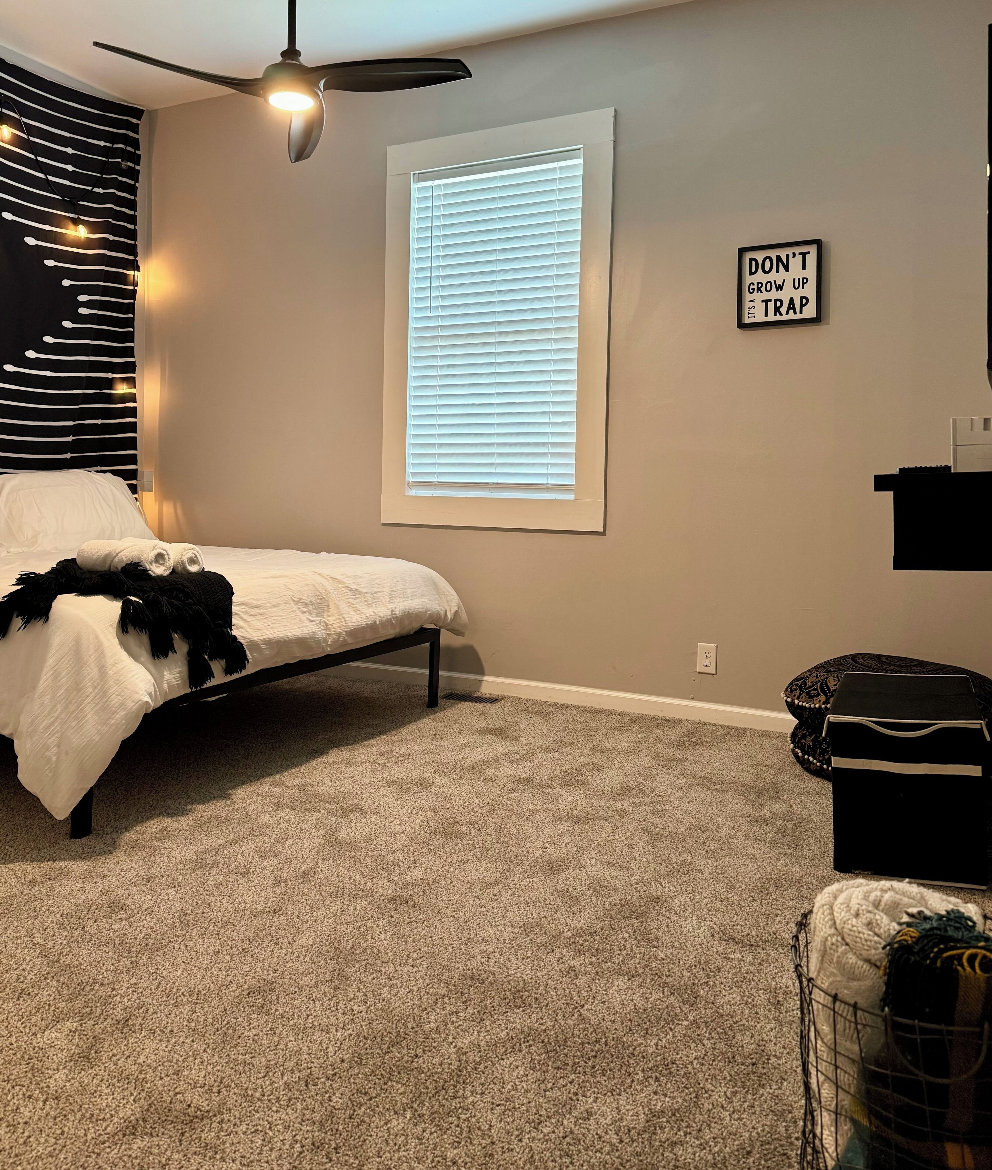 Une chambre avec un lit, un ventilateur de plafond, une fenêtre avec des stores, une citation inspirante sur le mur et une petite boîte de rangement.