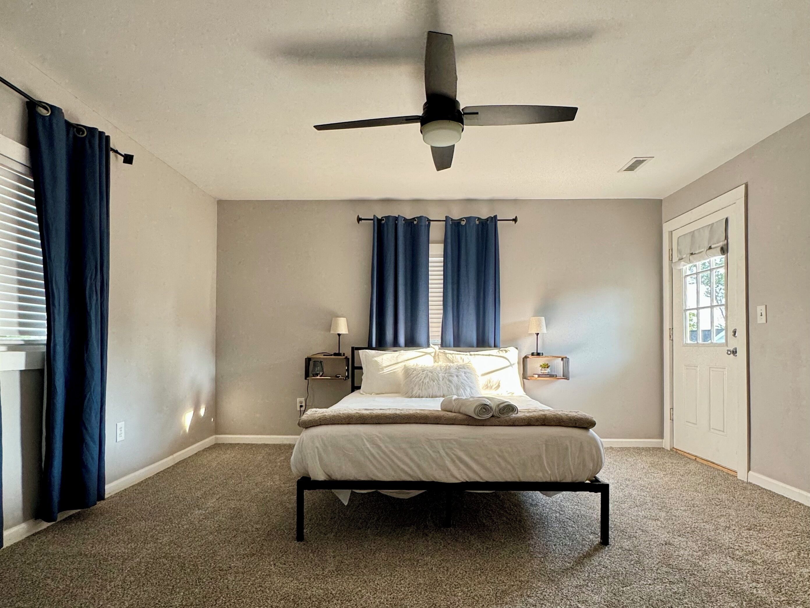Une chambre à coucher comprenant un lit, un ventilateur de plafond, une fenêtre avec des rideaux, une table de chevet avec une lampe et une porte.