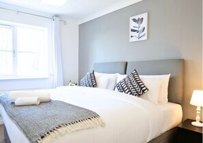 A neatly made bed with white and patterned pillows, a gray throw blanket, and a wall-mounted plant.