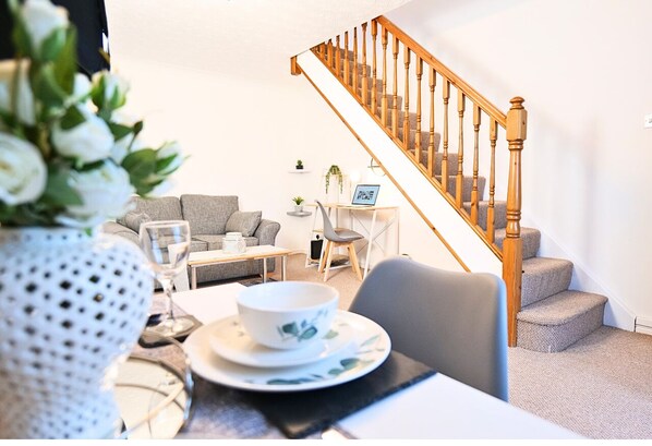 A living room with a staircase, a dining table set with plates and a bowl, and a vase with flowers.