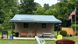 Un petit bâtiment d’un seul étage, avec un toit en métal bleu, une véranda avec des chaises en bois, une table et un drapeau.